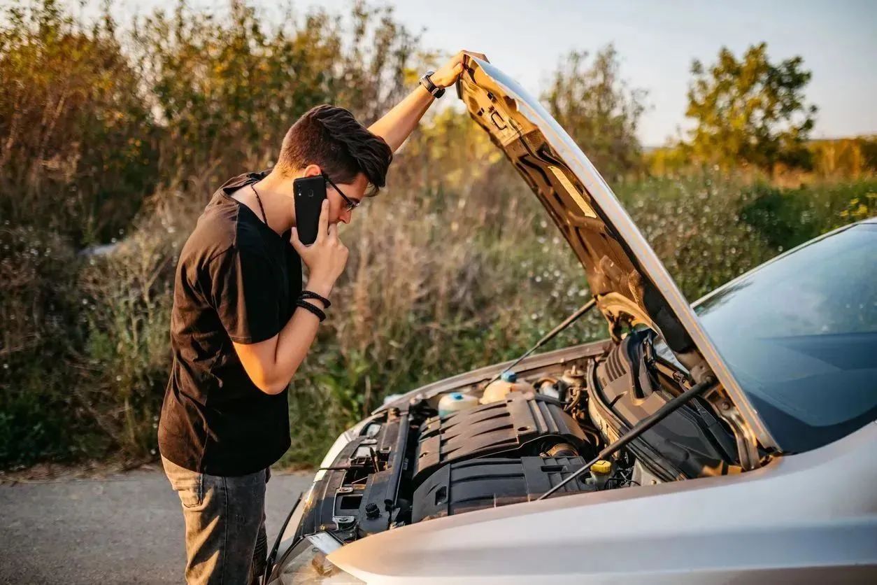 Driver looking at car engine while speaking on the phone in an outdoor setting.