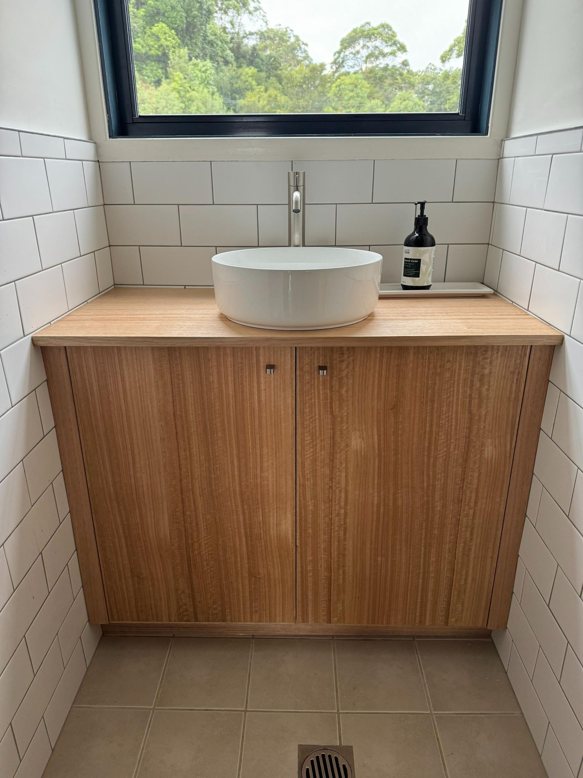 Bathroom with wood cabinet, white sink, and window overlooking trees. White tiled walls — BC Customs in Burrell Creek, NSW
