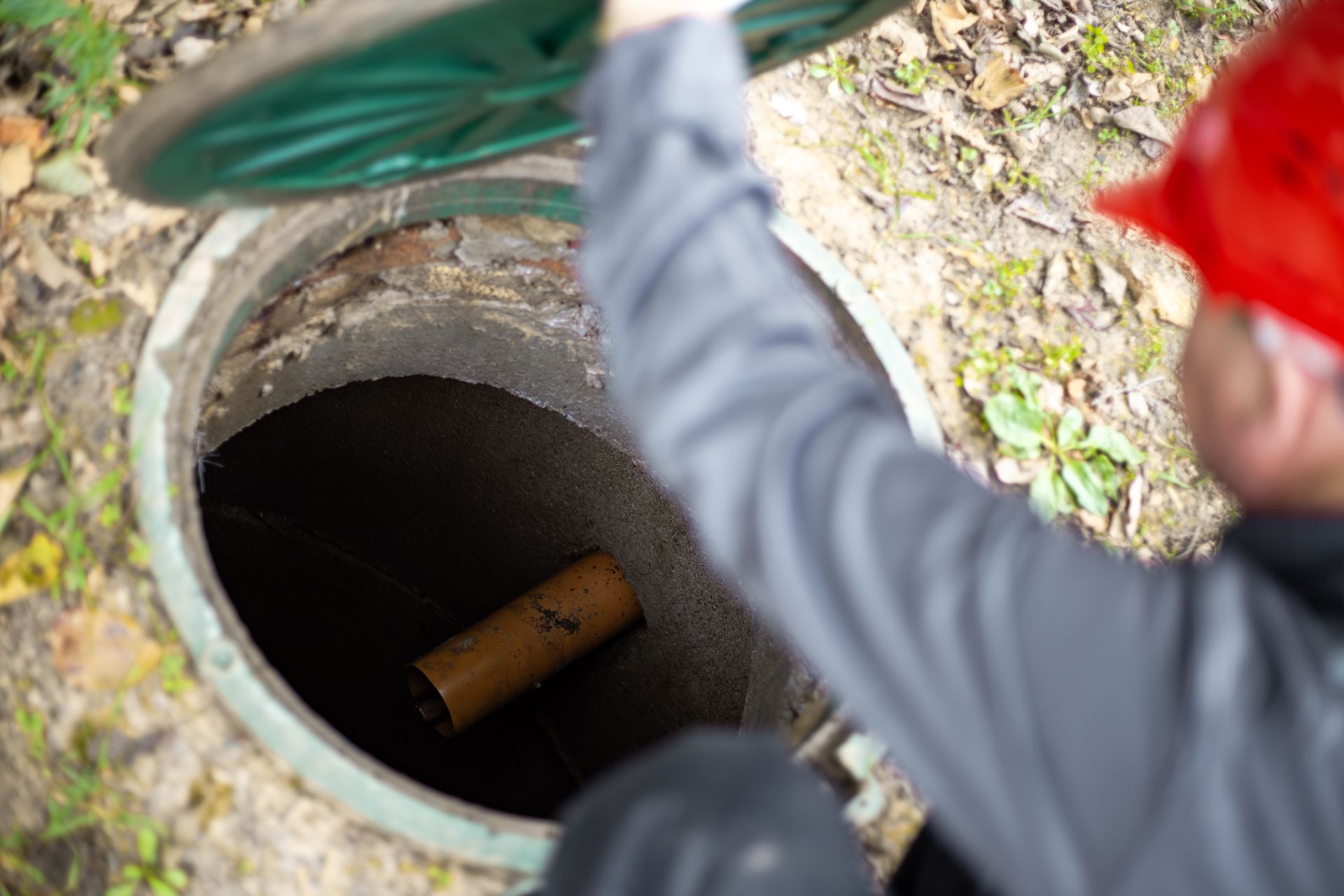 A worker looking into a manhole with a pipe inside, outdoors.