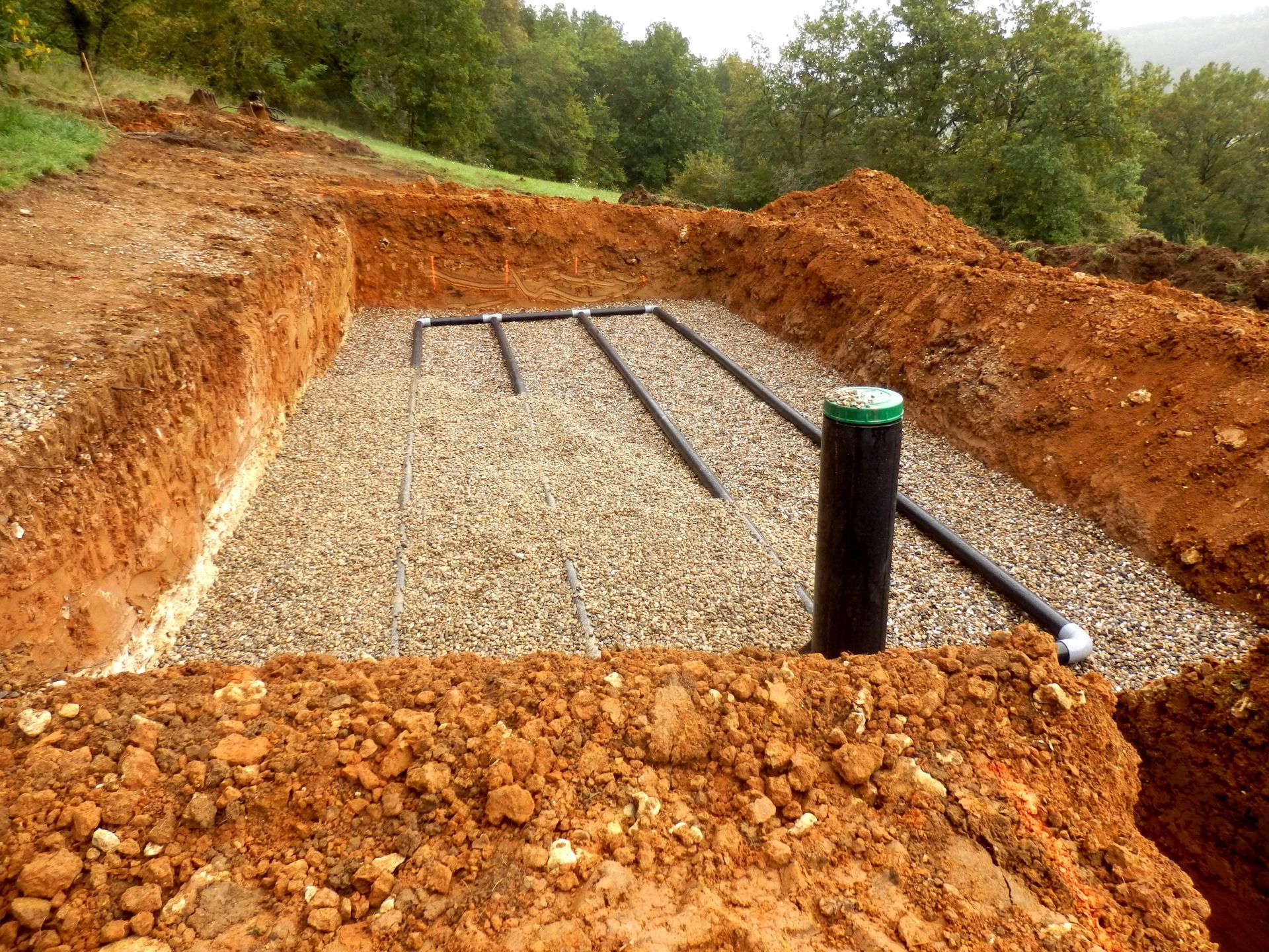 Excavated trench with gravel base and black pipes, surrounded by red soil and trees.