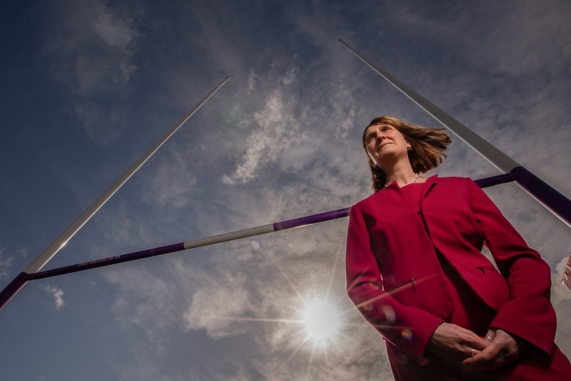 A woman in a red jacket is standing in front of a pole vault bar.