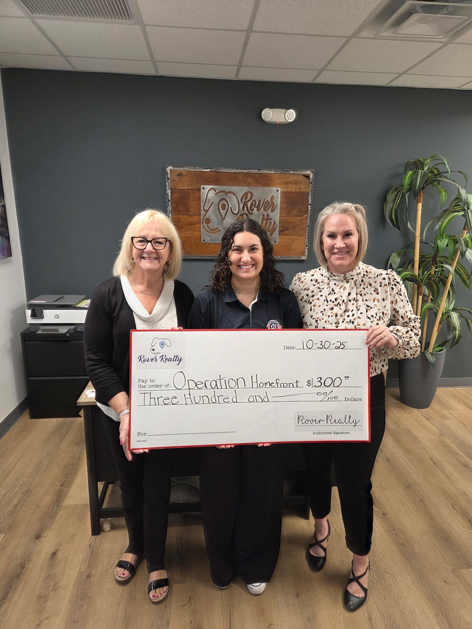 Three people standing indoors holding a large white sign in front of a dark wall and potted plant.