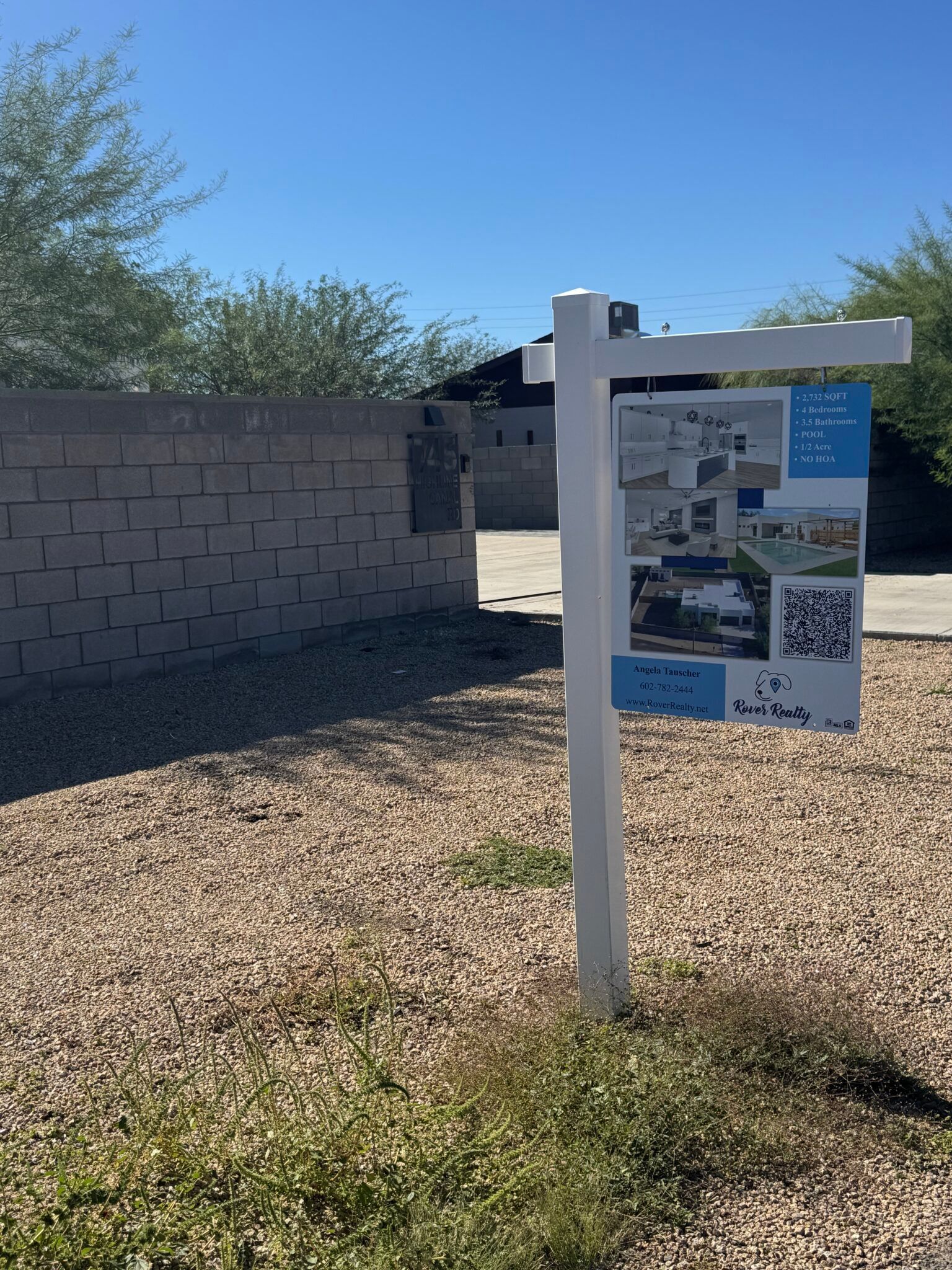 White signpost with a park information sign beside a gravel path, wall, and trees under a blue sky.