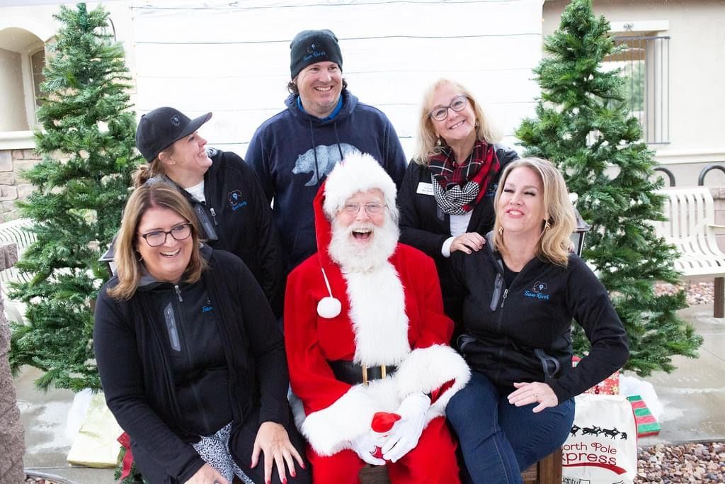 Santa seated with five people in winter clothes, posing in front of decorated Christmas trees.