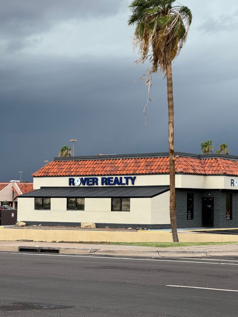 Corner business building with a red tile roof and sign reading “River Realty” under a cloudy sky.