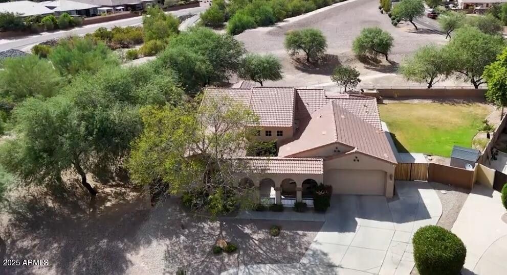 Aerial view of a tan stucco house with a tiled roof, driveway, and trees in a suburban neighborhood