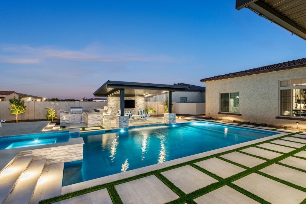 Modern backyard pool at dusk with glowing water, stone patio, and contemporary house exterior