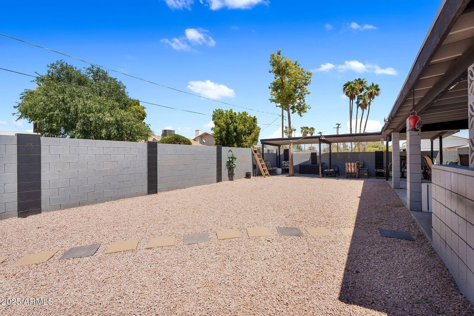 Spacious gravel backyard with a covered patio, gray fence, and tall palm trees in bright sunlight