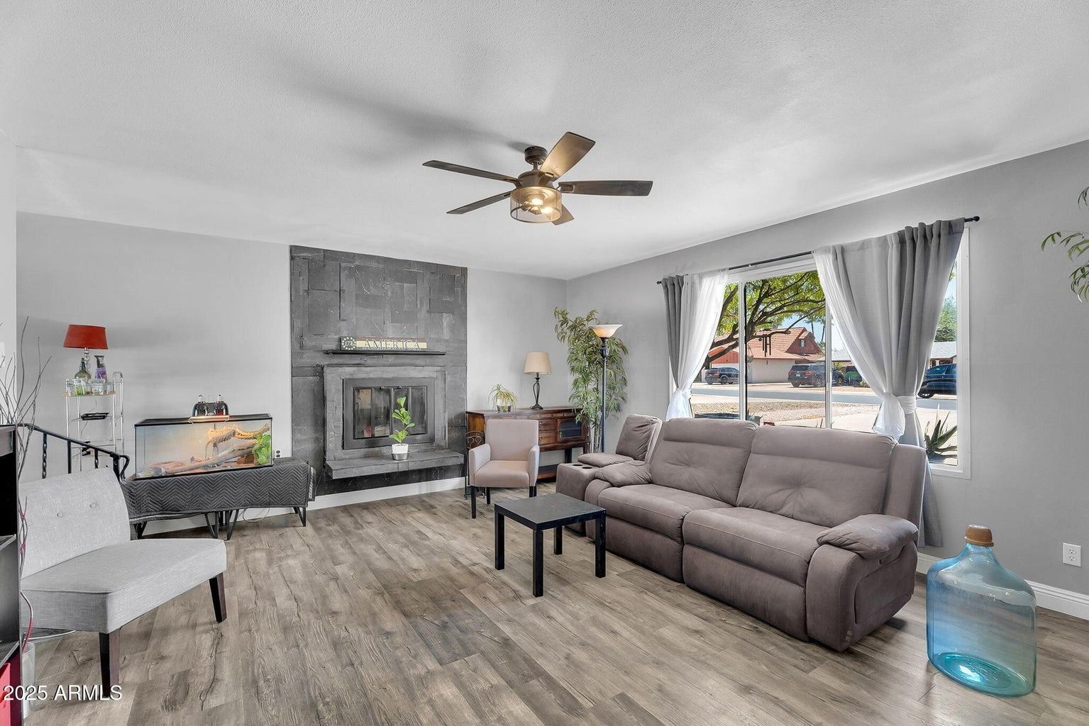 Bright living room with gray sofa, fireplace, large window, and ceiling fan on wood-look flooring.