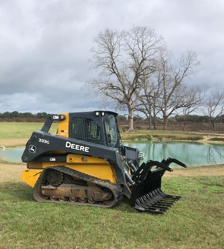John Deere 333G compact track loader with grapple bucket on grassy bank, by a pond. Overcast sky.