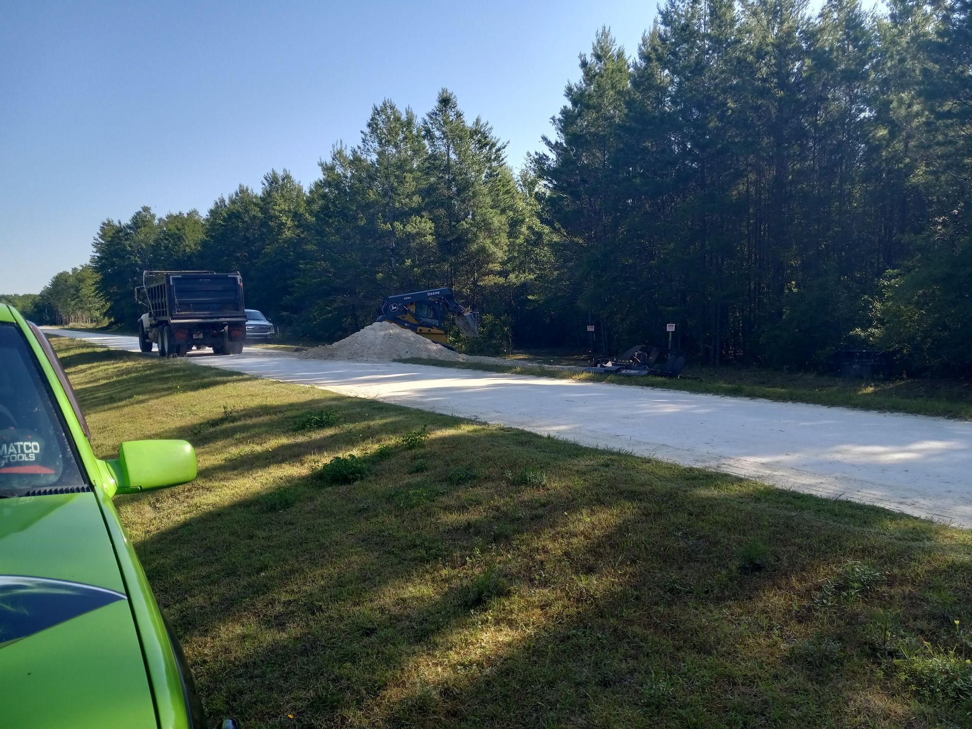 Green car parked on grass, dump truck on gravel road next to a pile of material, trees in background.
