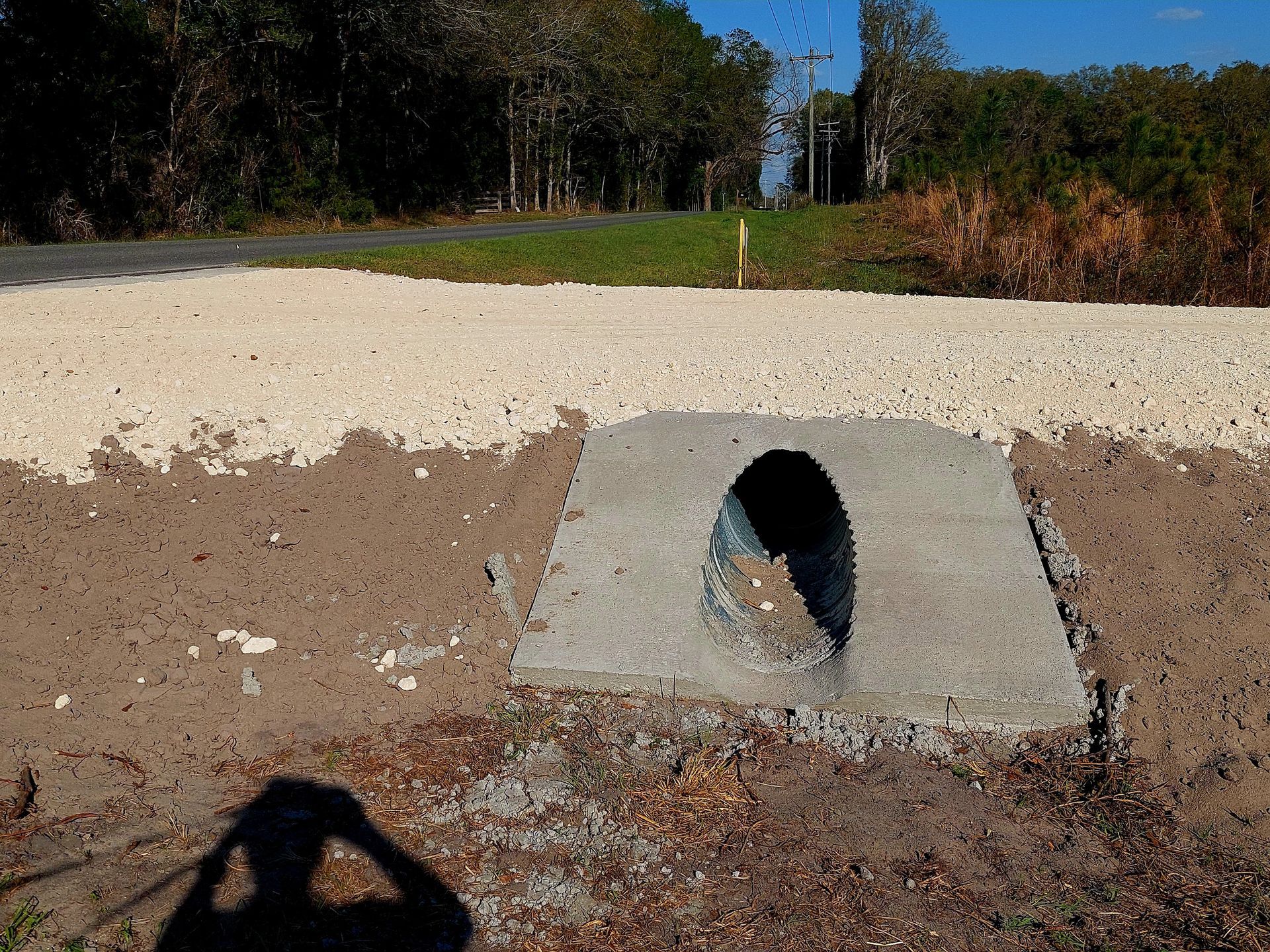 Concrete culvert under a road with gravel shoulder, leading to a grassy area and trees.