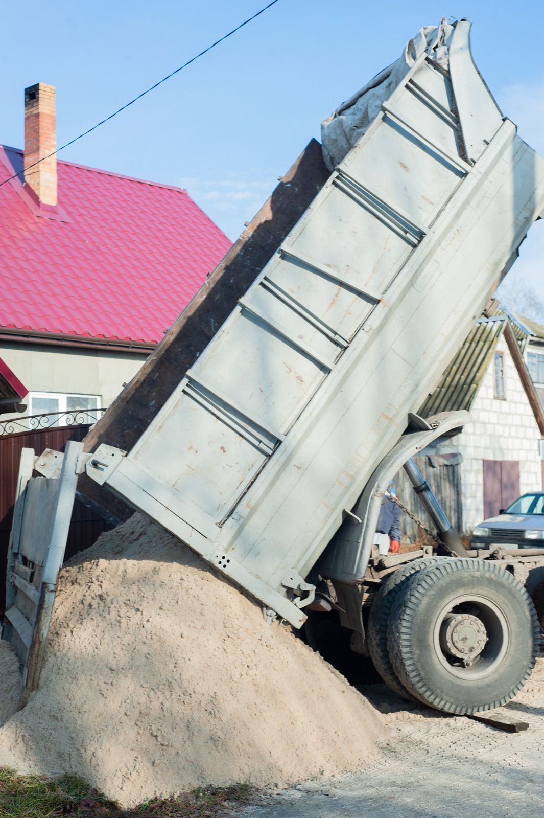 Dump truck tilted, unloading sand near a house with red roof.