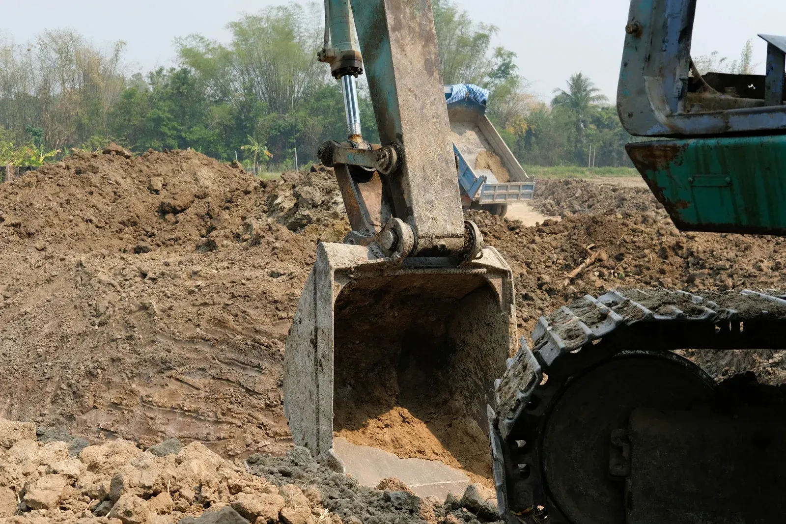 Excavator bucket scooping up soil at a construction site; blue, green, and brown tones.