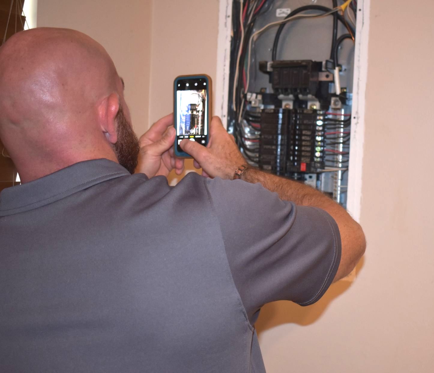 Man taking a photo of an electrical panel with a smartphone; beige wall in the background.
