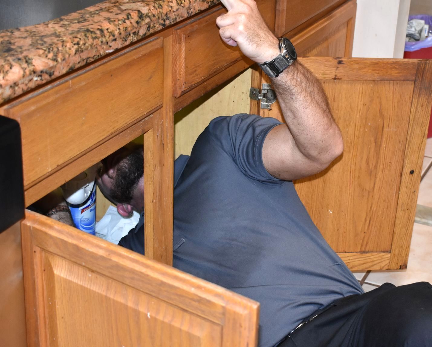 Person working under a kitchen sink. Wooden cabinet with open door.