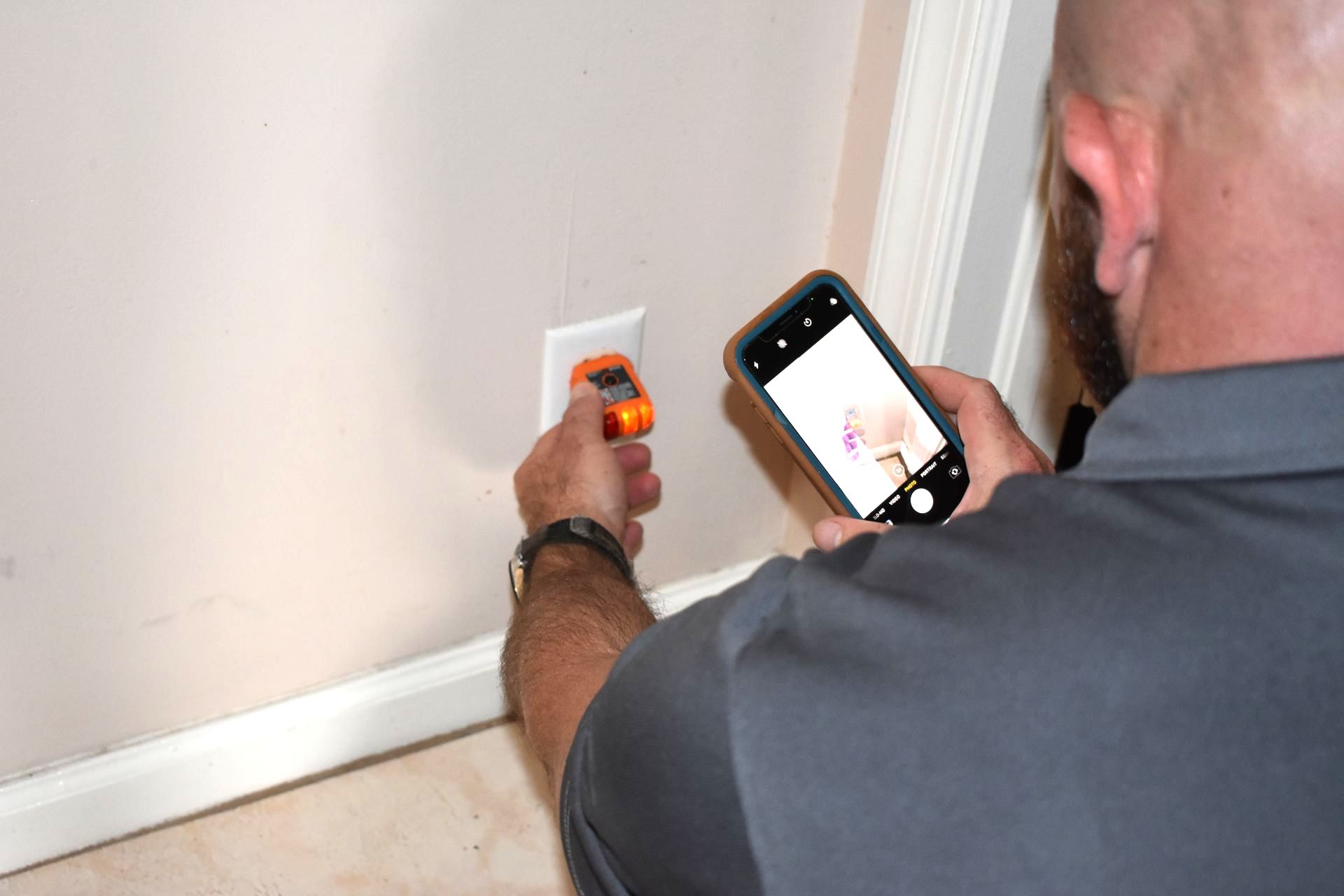 Man using a multimeter on an outlet while taking a photo with his smartphone. Beige wall, white trim.