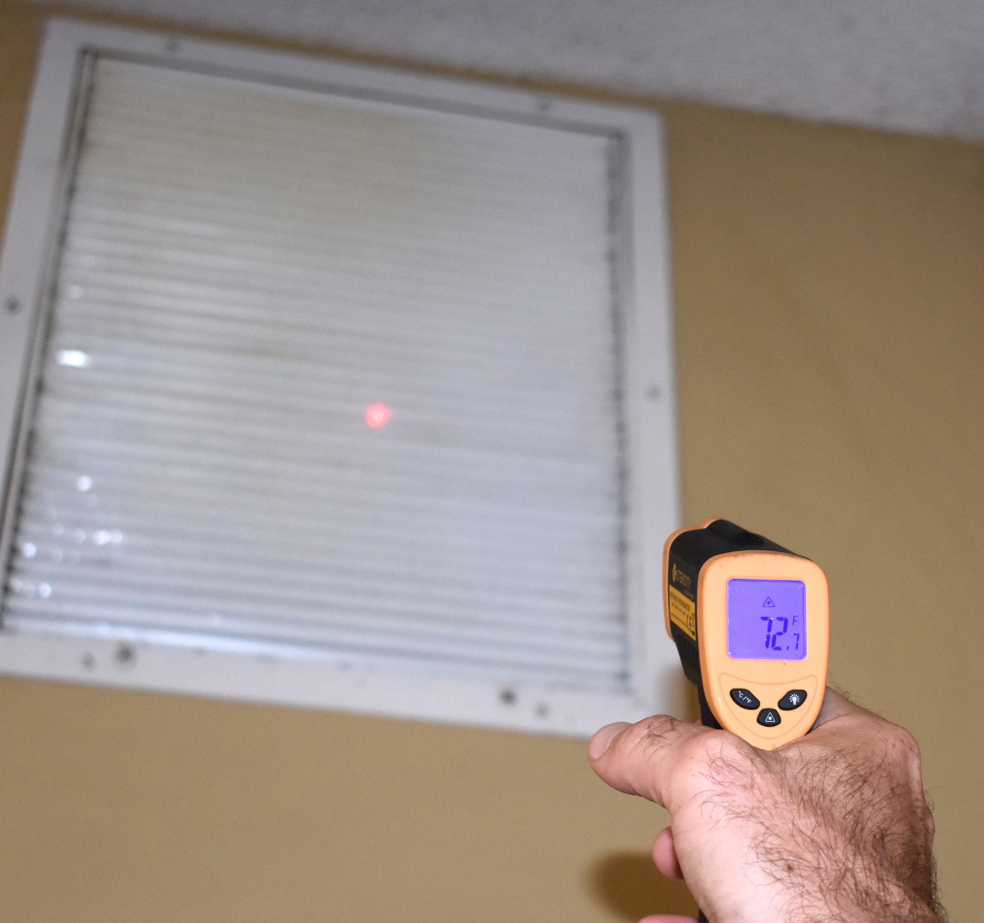 Hand holding a temperature gun pointing at a white vent with a red laser dot; the screen displays 72.7.