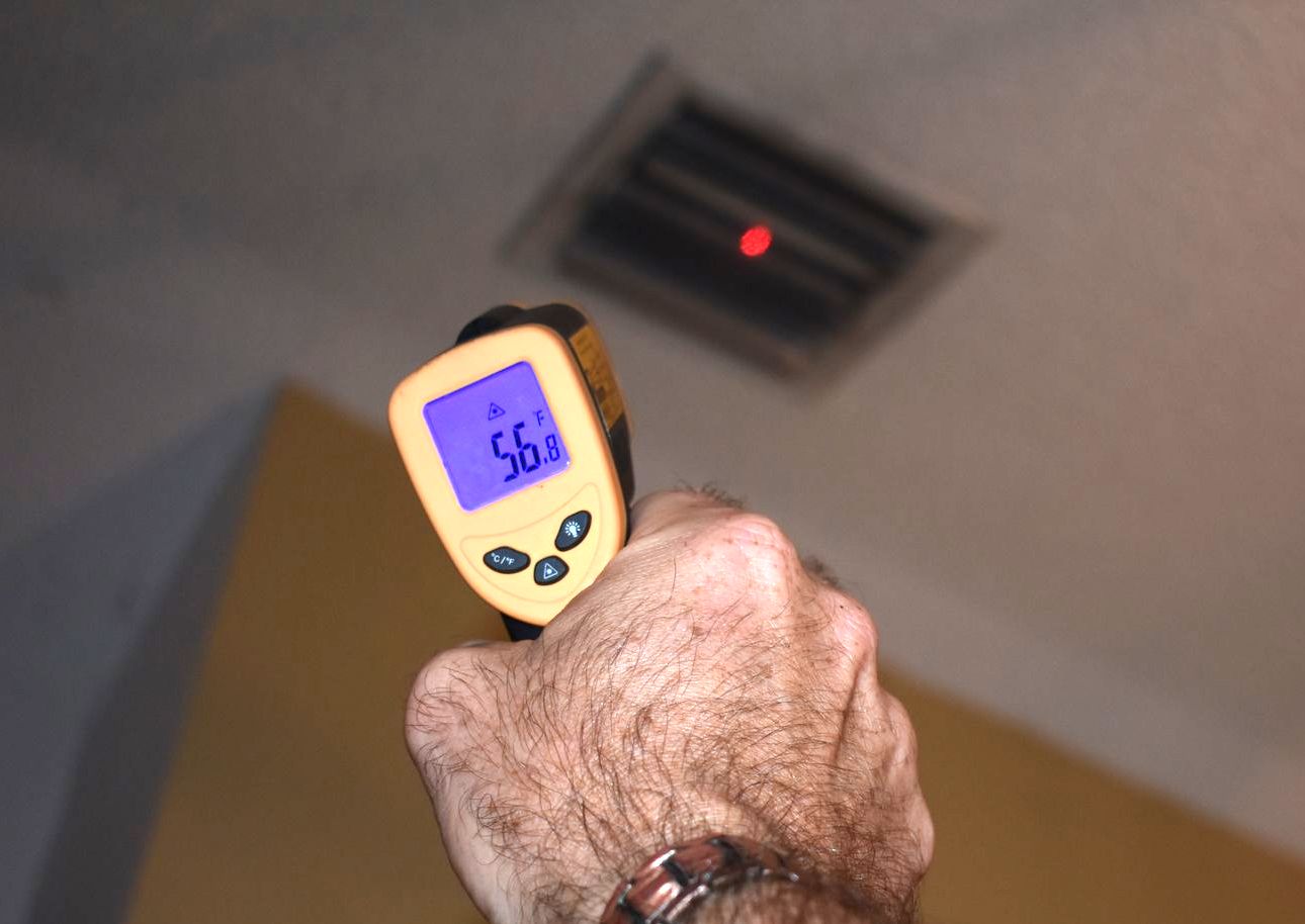 Hand holding a yellow infrared thermometer pointed at an air vent in a ceiling; the device reads 56 degrees.