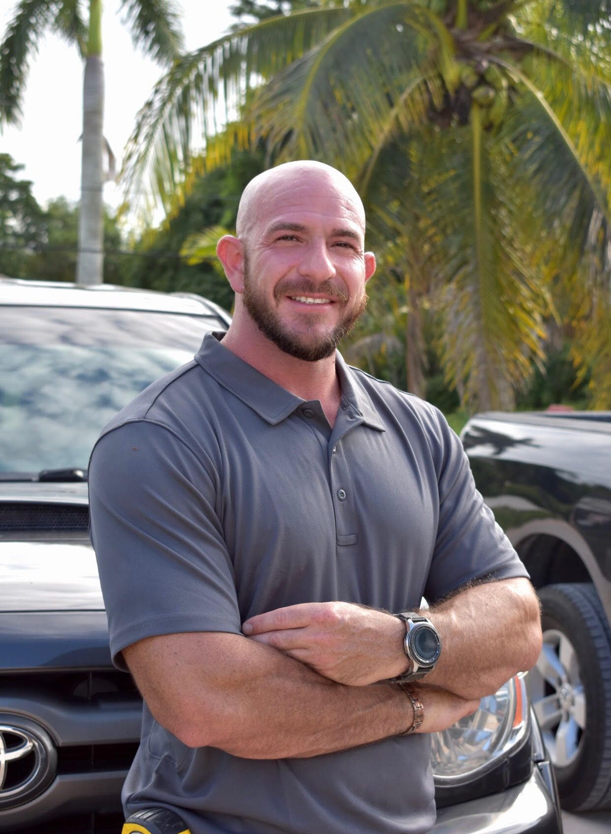 Man with short hair and beard smiles, arms crossed, standing in front of a black vehicle.