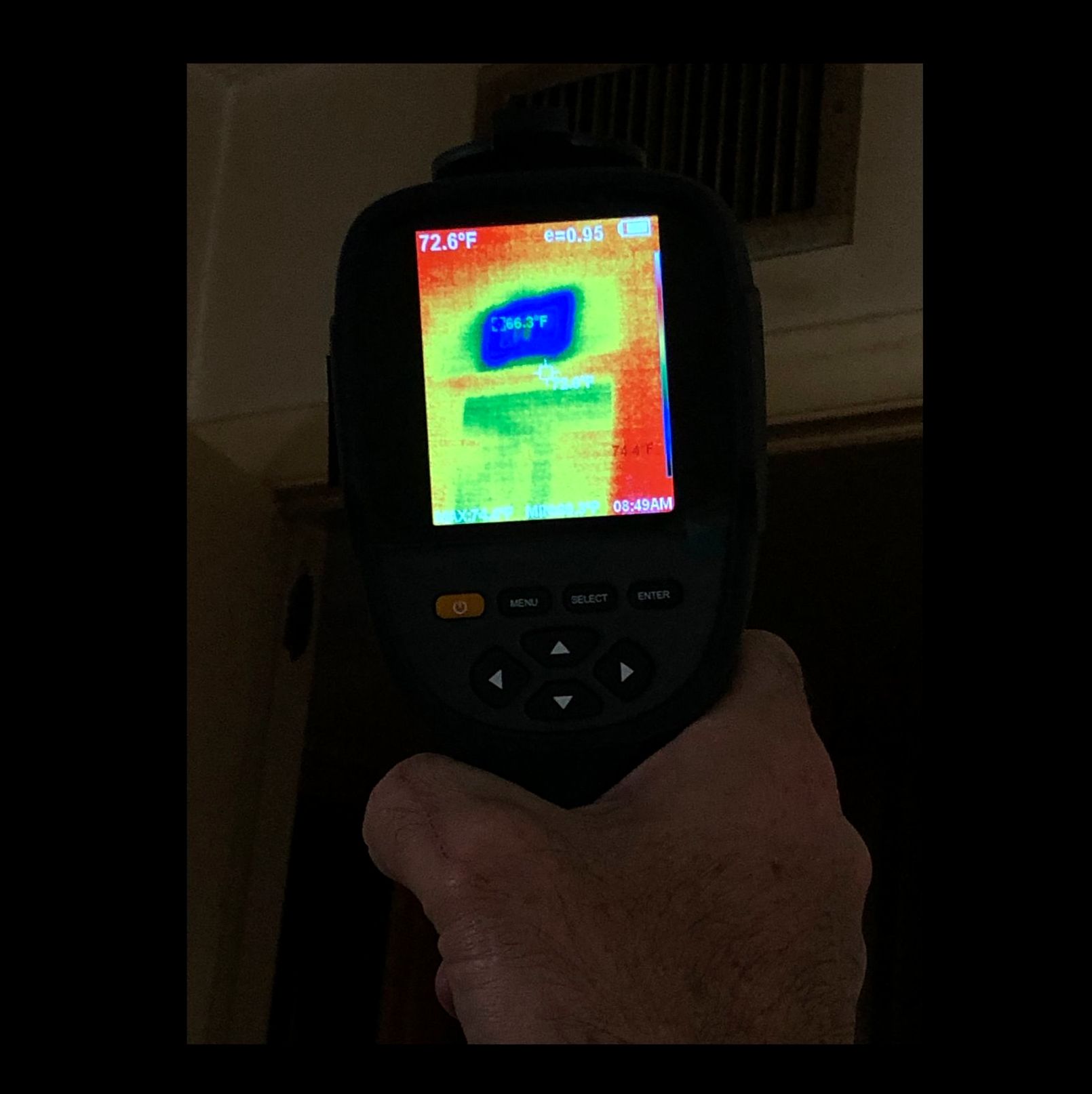 Hand holding a thermal imaging camera pointed at a wall. The screen shows temperature variations in various colors.