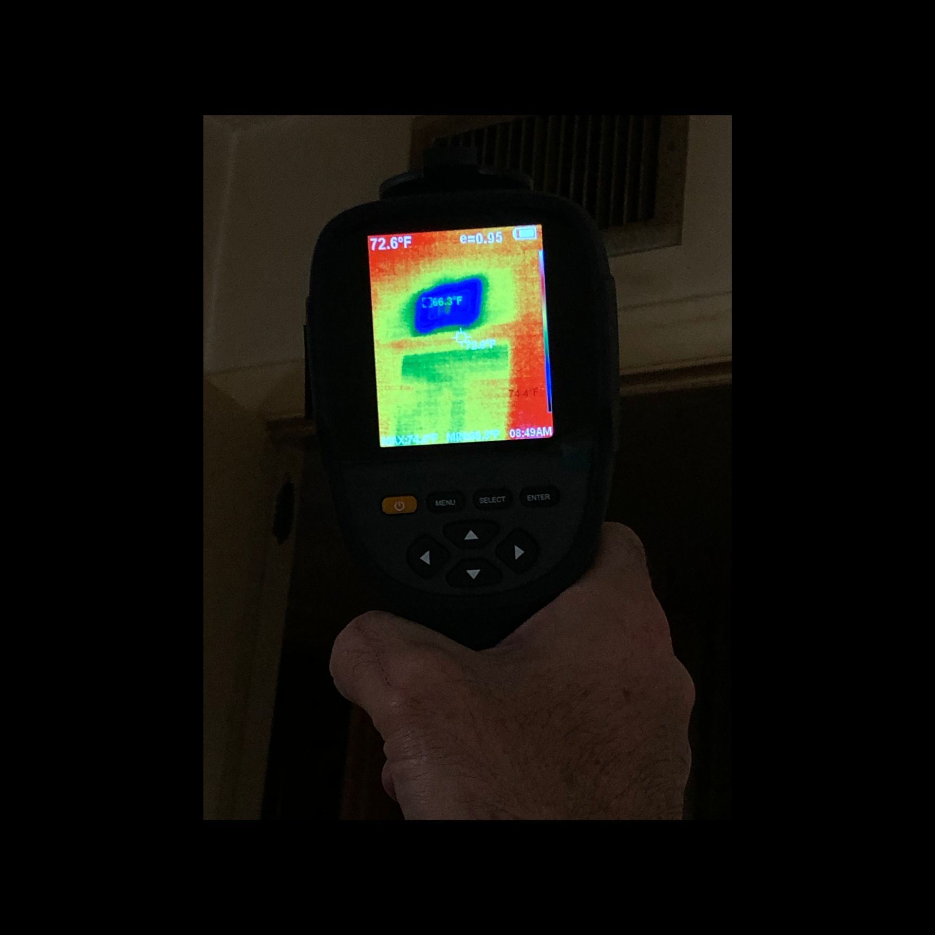 A hand holding a thermal imaging camera pointed at a vent. The camera displays heat readings in varying colors.