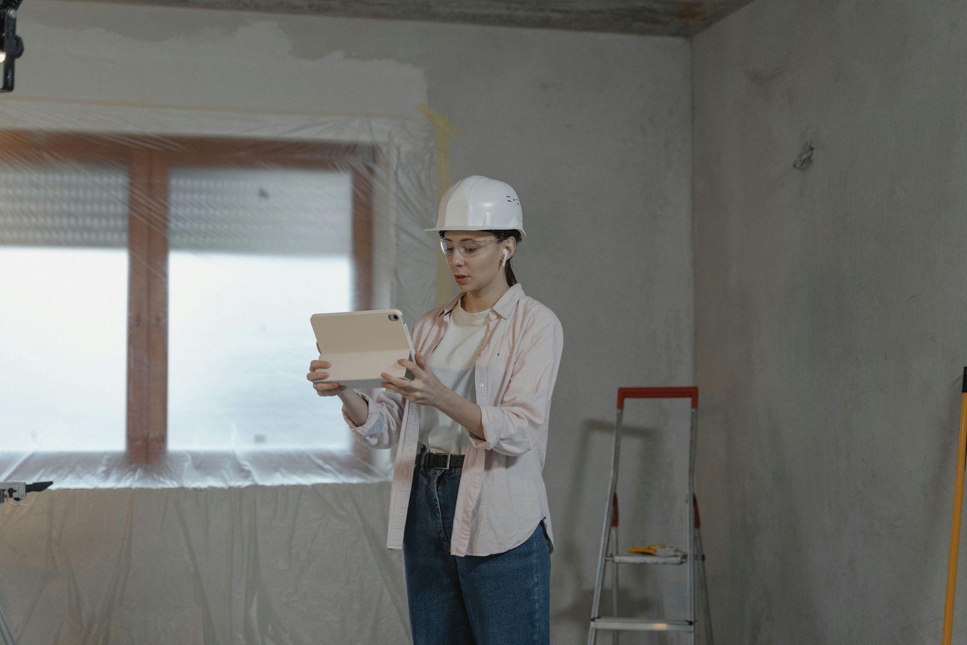 Woman in hard hat, shirt, and jeans holding a tablet, inspecting a room under construction.