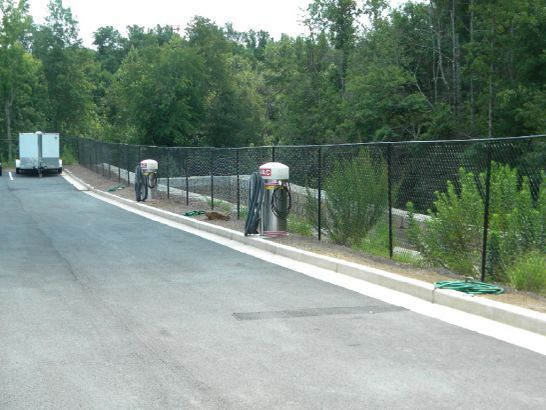 Workers near a black chain-link fence along a road, with green foliage and a trailer in the background.