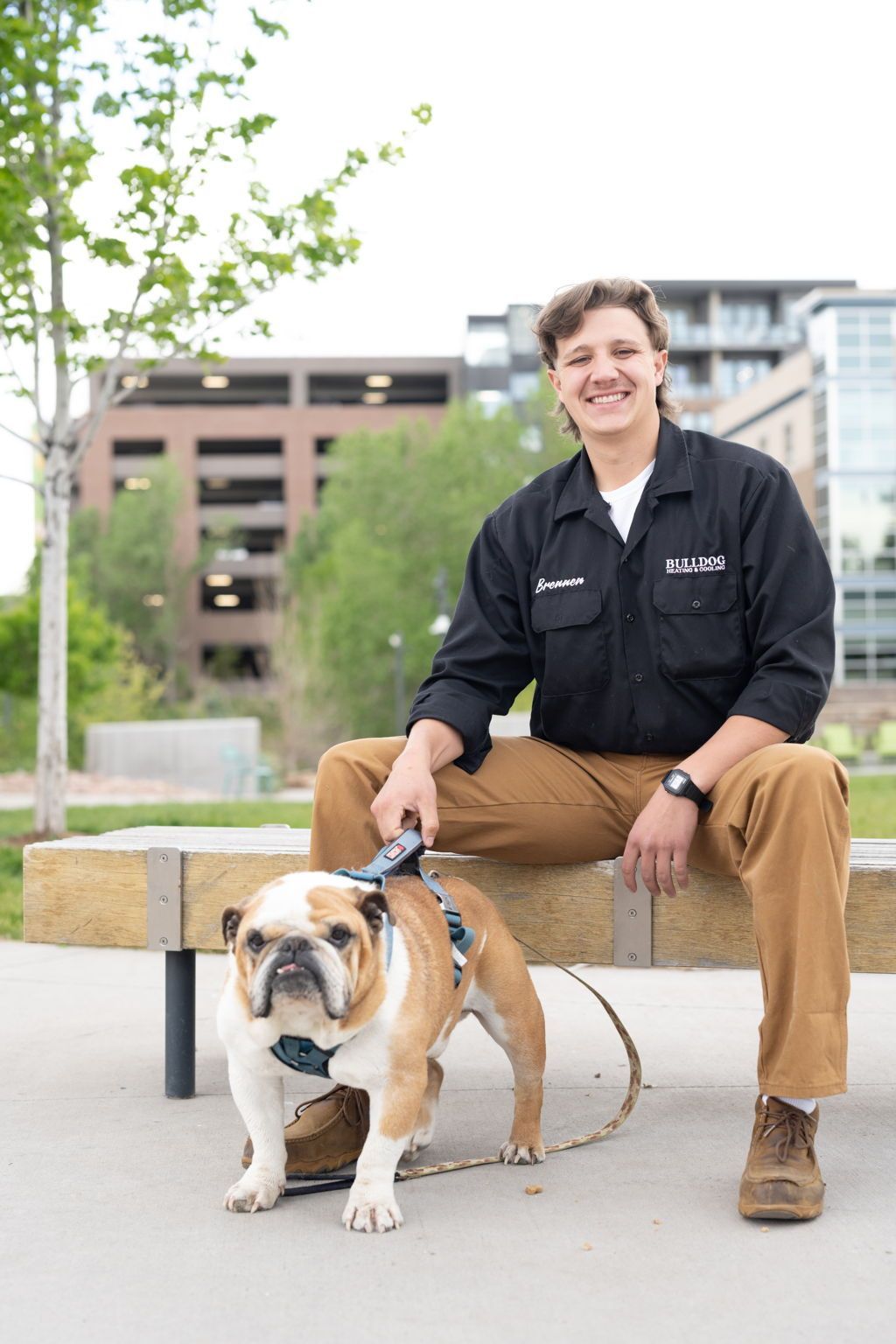 A smiling person in a black shirt and tan pants sits on a wooden bench outdoors next to a bulldog wearing a grey harness.