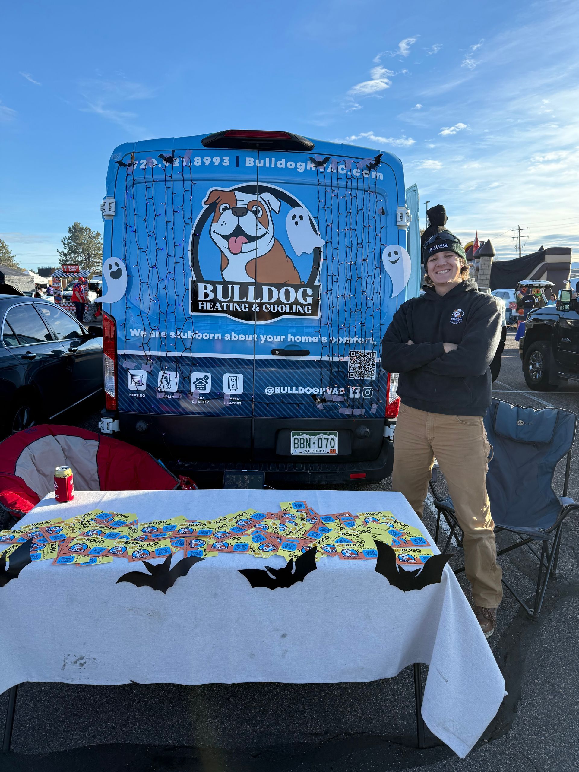 A person stands with arms crossed in front of a blue Bulldog Plumbing van decorated for Halloween with a candy table.