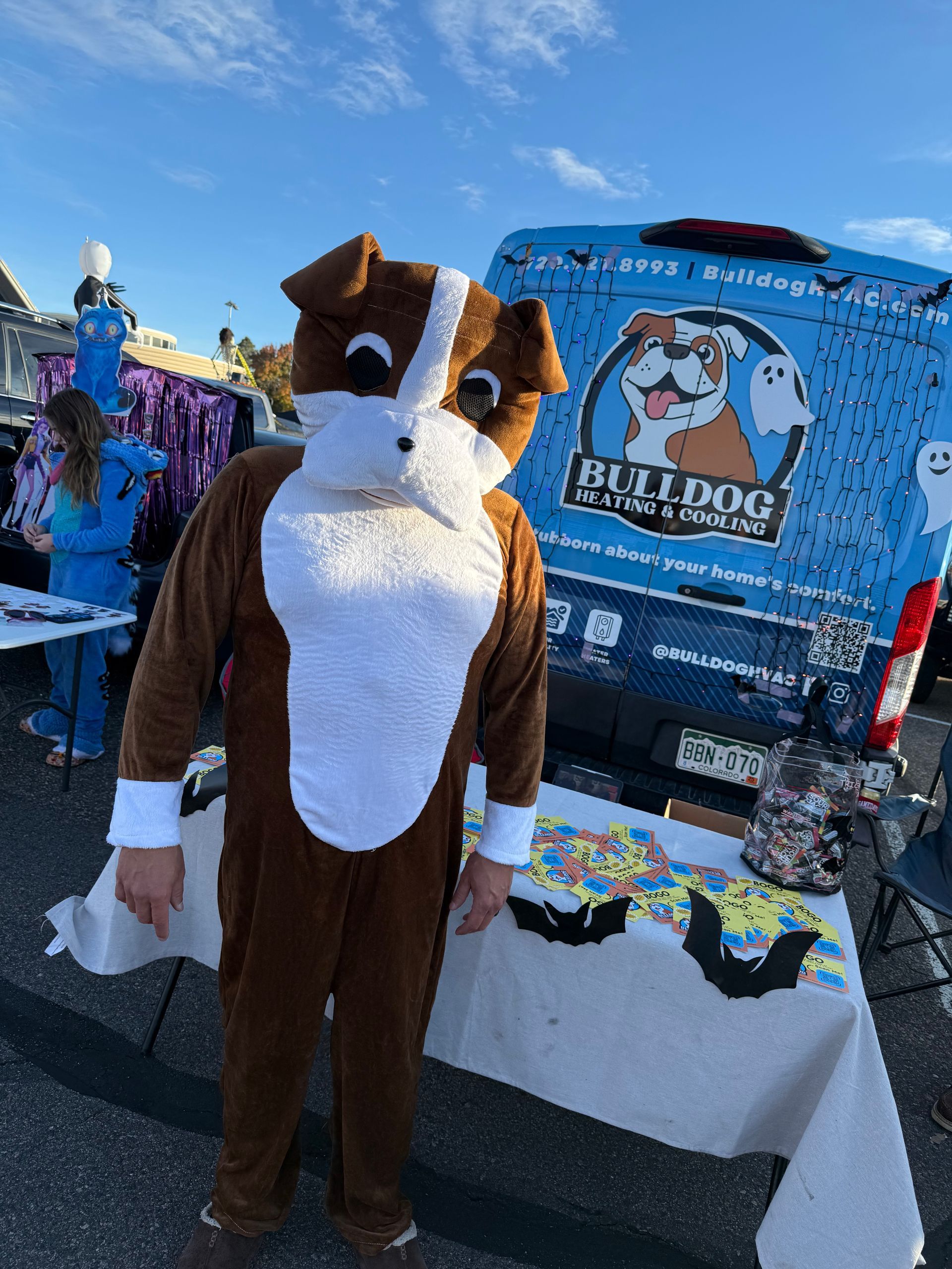 A person in a brown and white dog mascot costume stands in front of a blue Bulldog Moving & Storage van at an event.