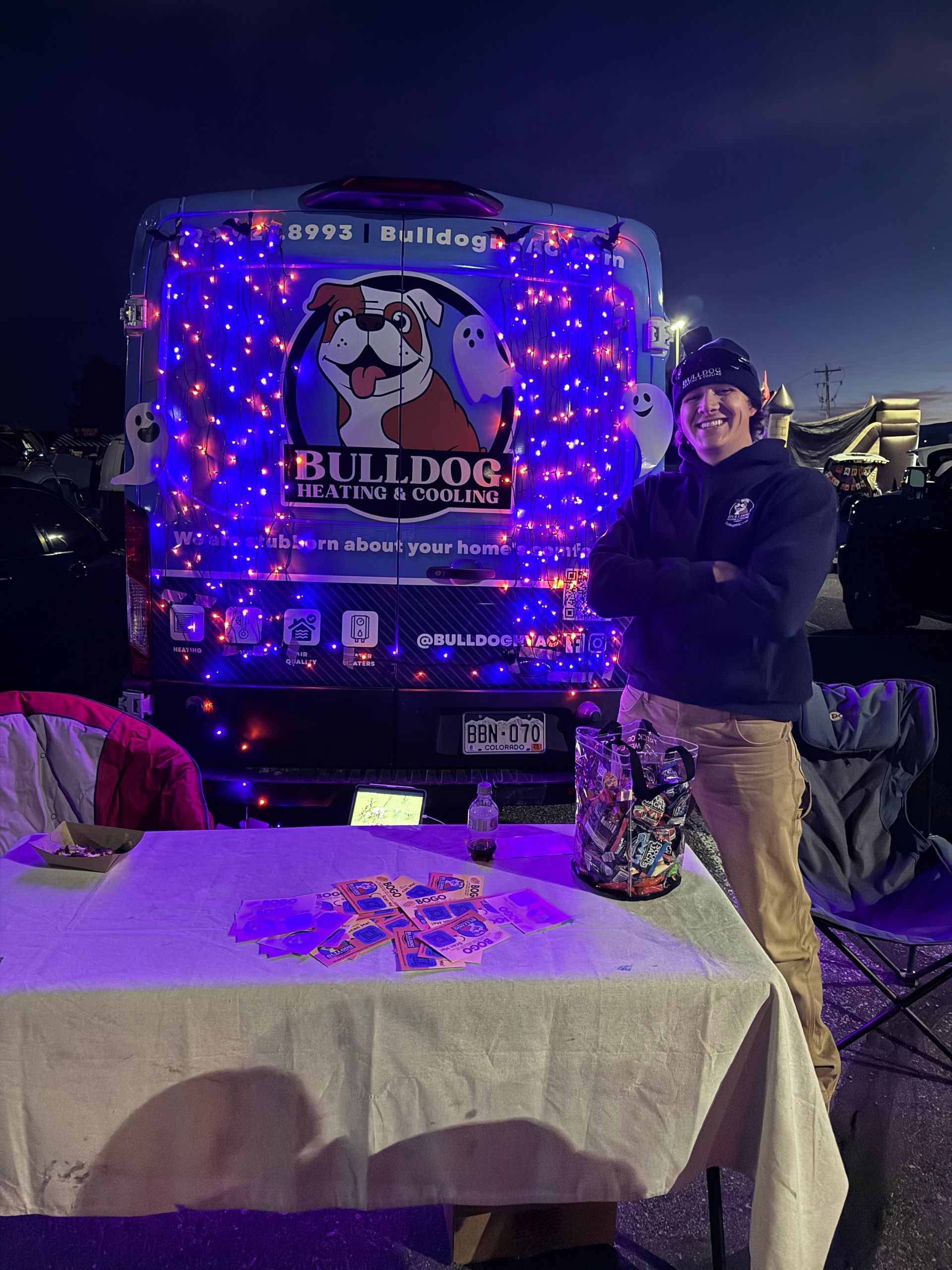 A person stands with arms crossed next to a van decorated with purple string lights and a Bulldog logo at night.