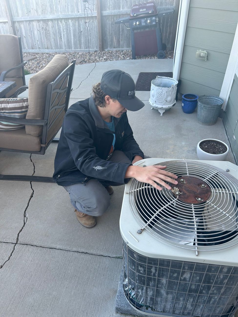 Person in a black jacket and cap inspects an AC unit outside.