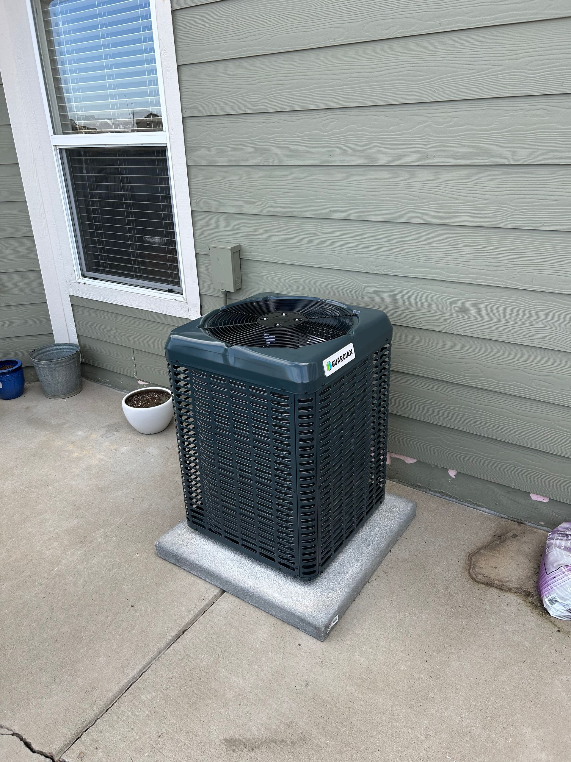 A black square HVAC outdoor unit sits on a concrete pad next to the light green siding and window of a house.