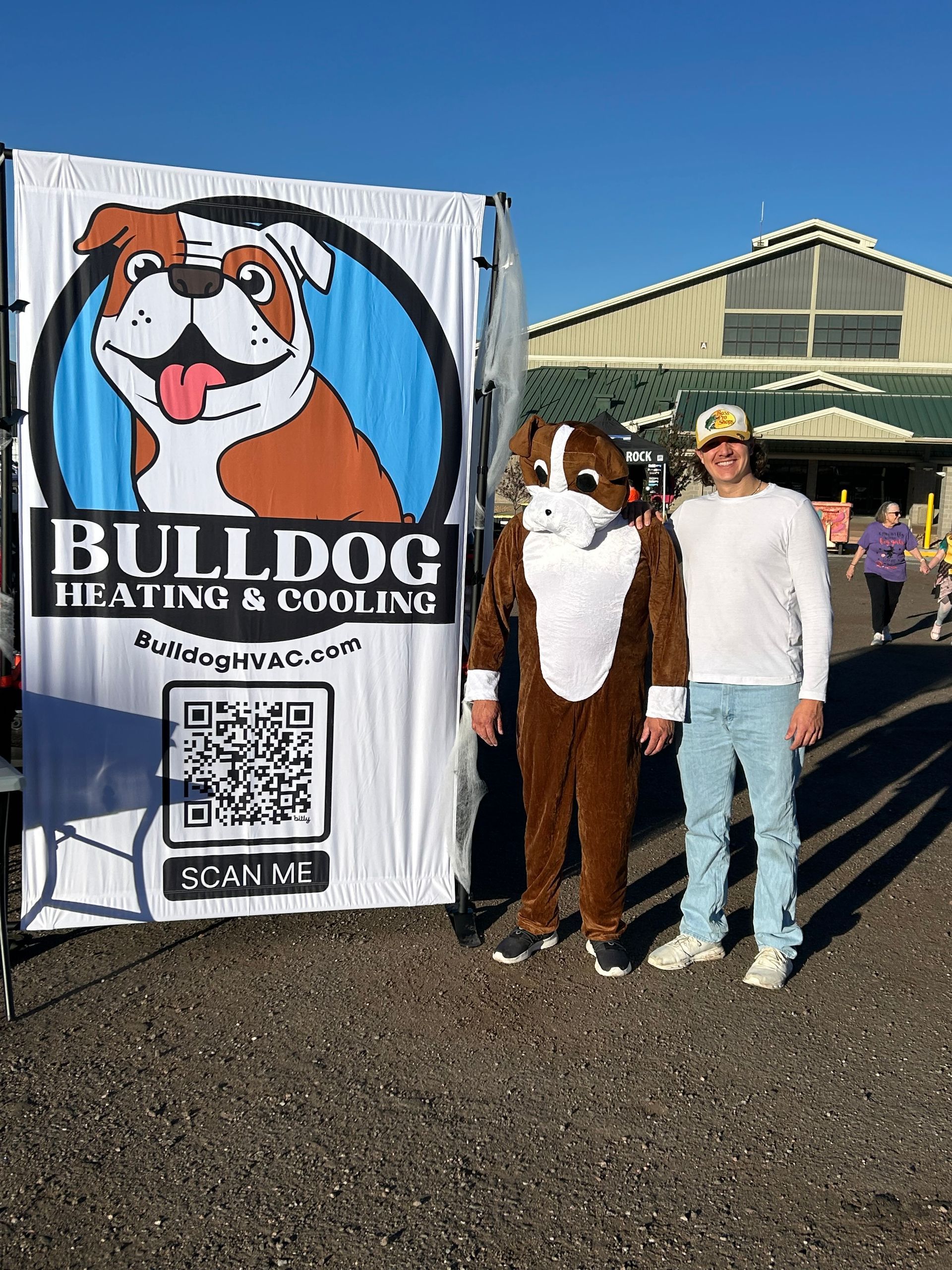A mascot in a bulldog costume and a person stand beside a Bulldog Heating & Cooling banner with a QR code outdoors.