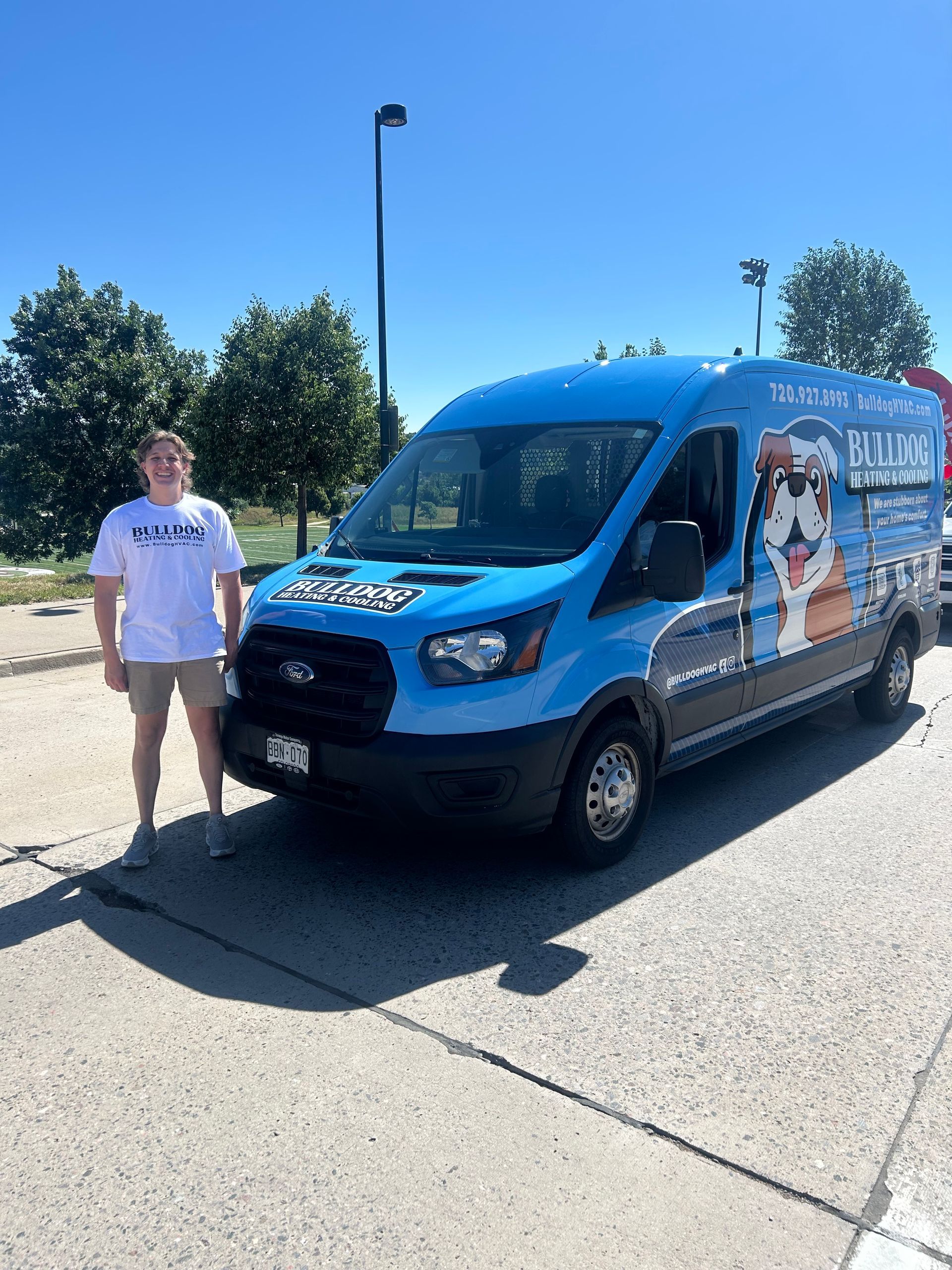 A person stands next to a bright blue van featuring a large bulldog logo, parked on a sunny paved lot.