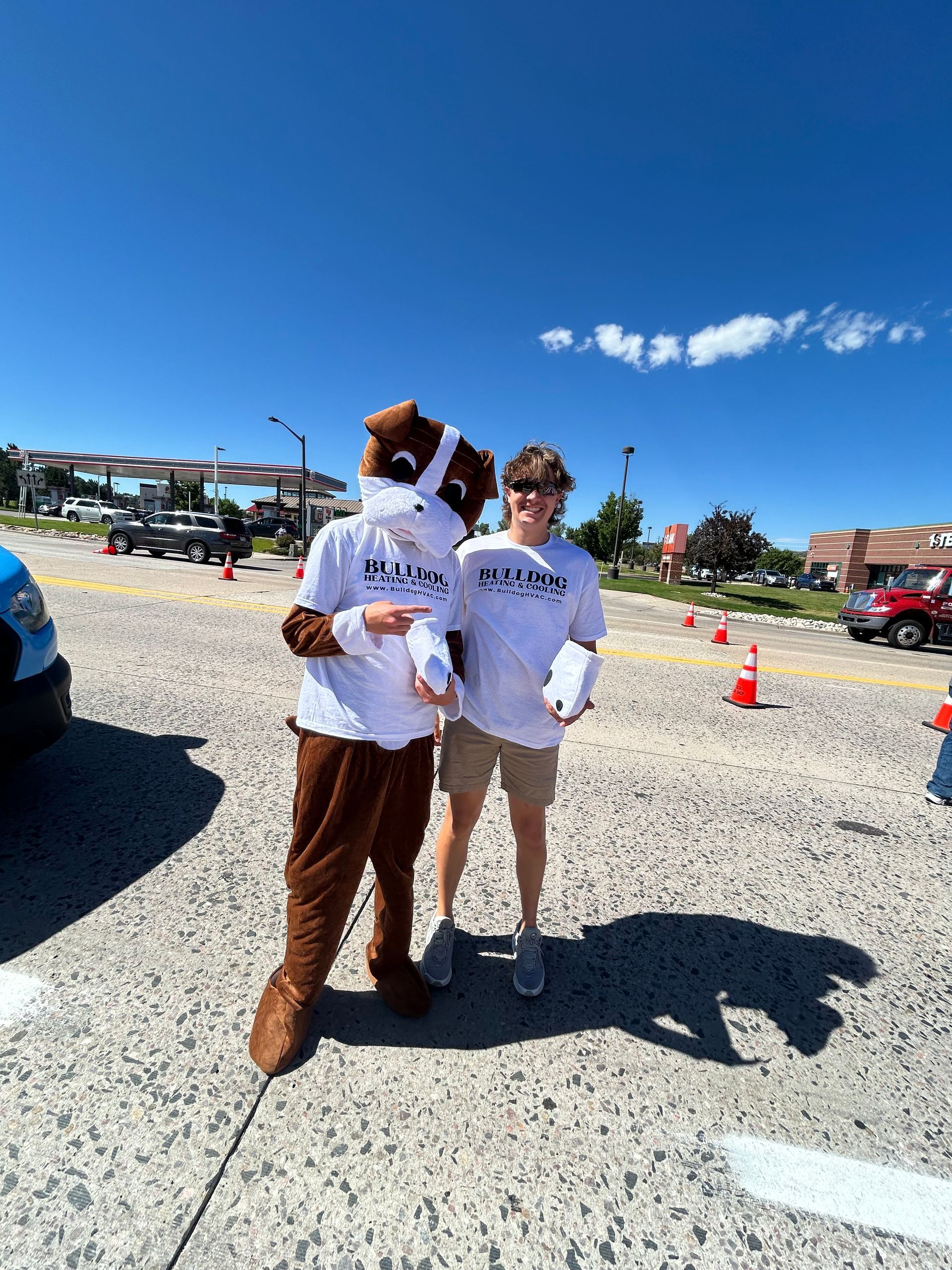 A person in a brown dog mascot costume stands next to a person in a white t-shirt in a sunny, gravel-covered parking lot.