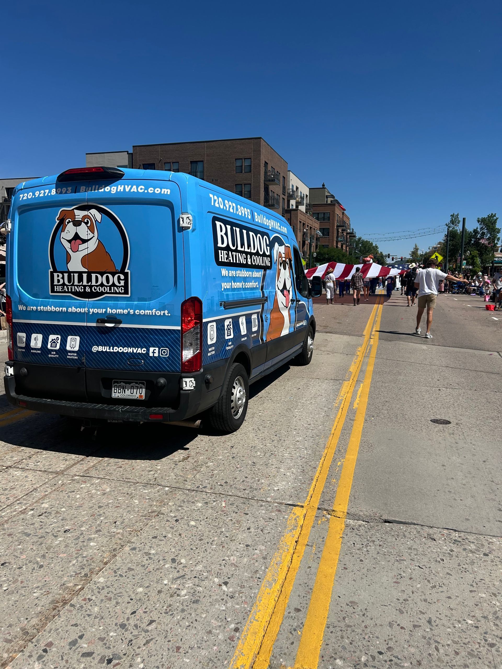 A blue Bulldog Plumbing van drives down a sunny street during a neighborhood parade.