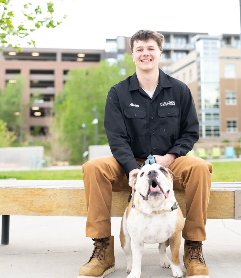 A smiling person sits on an outdoor bench with a bulldog standing between their legs against a blurred city background.