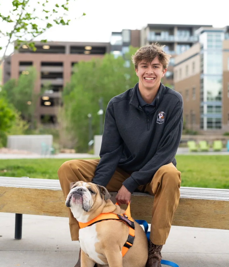 A smiling person sits on a wooden bench outdoors with a bulldog wearing an orange harness.