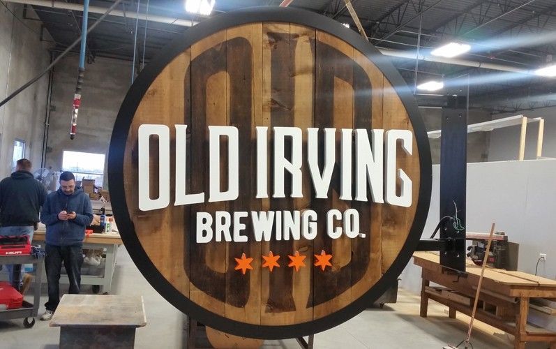 Old Irving Brewing Co. sign in workshop, circular wooden barrel shape with white lettering, orange stars, and people working.