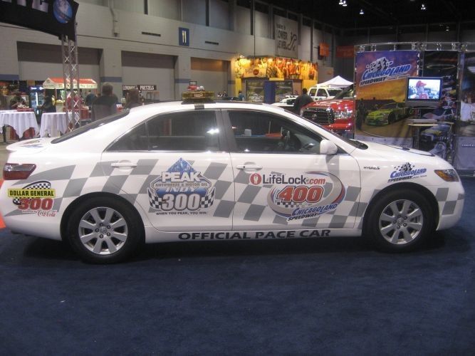 White Toyota Camry pace car with racing logos and checkered flag pattern.