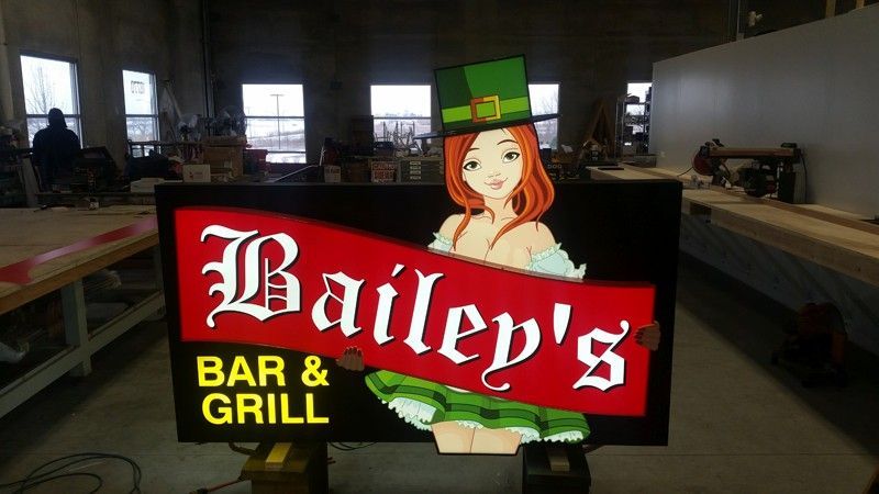 Bailey's Bar & Grill sign with cartoon woman in a green leprechaun hat and outfit. Red banner across text.