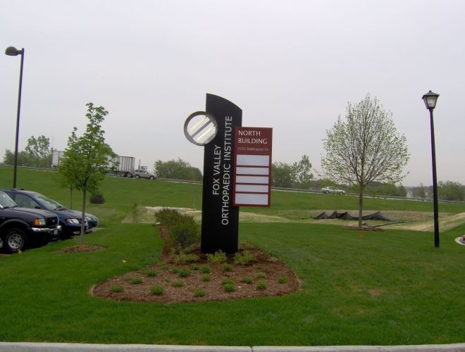 Sign for Fox Valley Orthopedic Institute; lists building name & addresses in landscaped area with parked cars.
