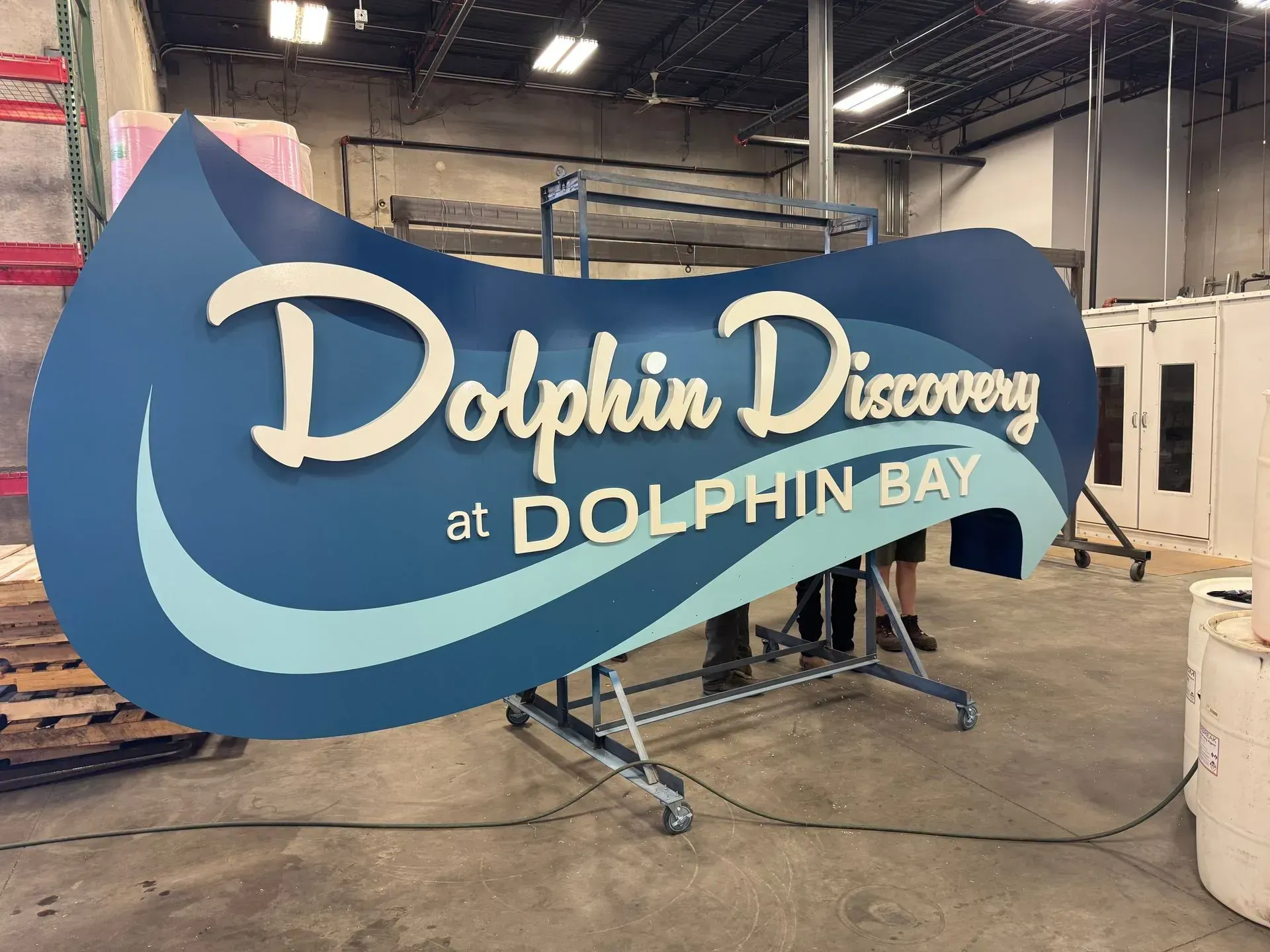 Blue “Dolphin Discovery at Dolphin Bay” sign in an indoor workshop or warehouse setting.
