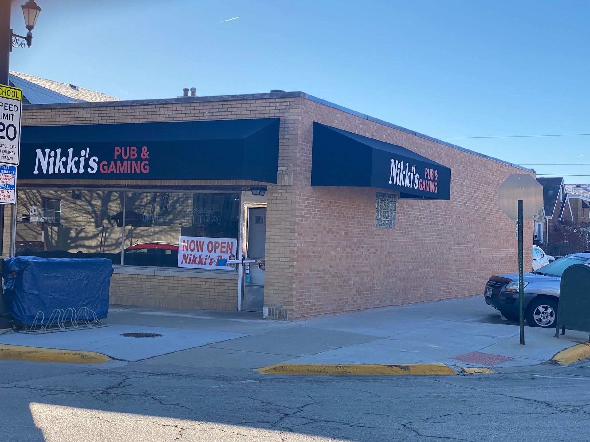 Nikki's Pub & Gaming, brick building with black awning and sign. Door open sign visible. Street view with blue bin and parked car.