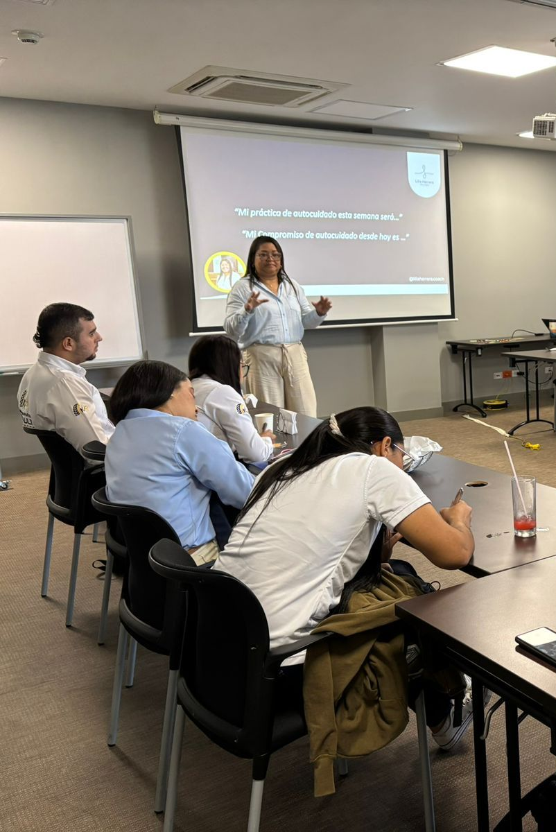 Una mujer da una presentación en una sala de reuniones con una diapositiva proyectada. Los asistentes escuchan atentamente.