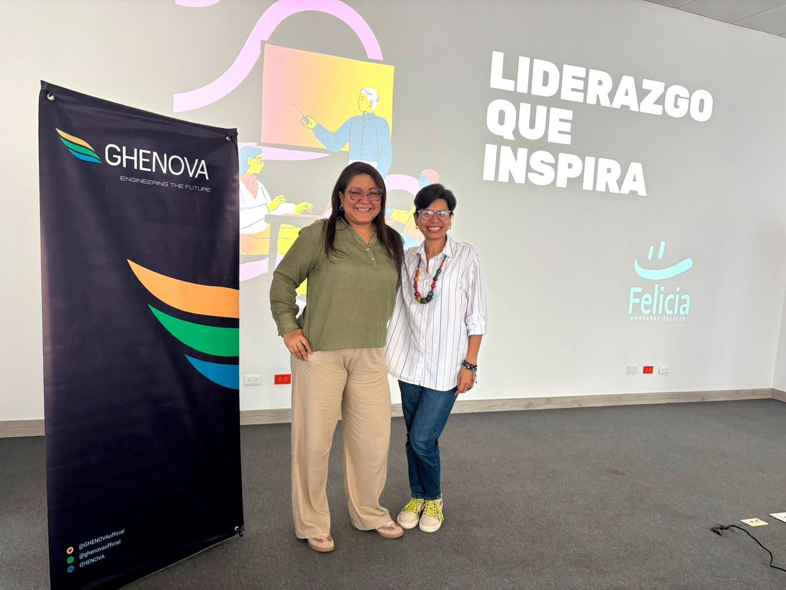 Dos mujeres están de pie junto a una pancarta. Una lleva una camisa verde y pantalones color canela; l