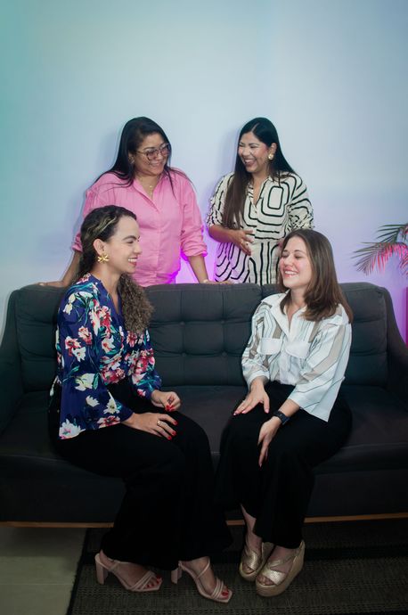Cuatro mujeres conversando, dos sentadas en un sofá y dos de pie detrás. Ropa rosa, negra y blanca.