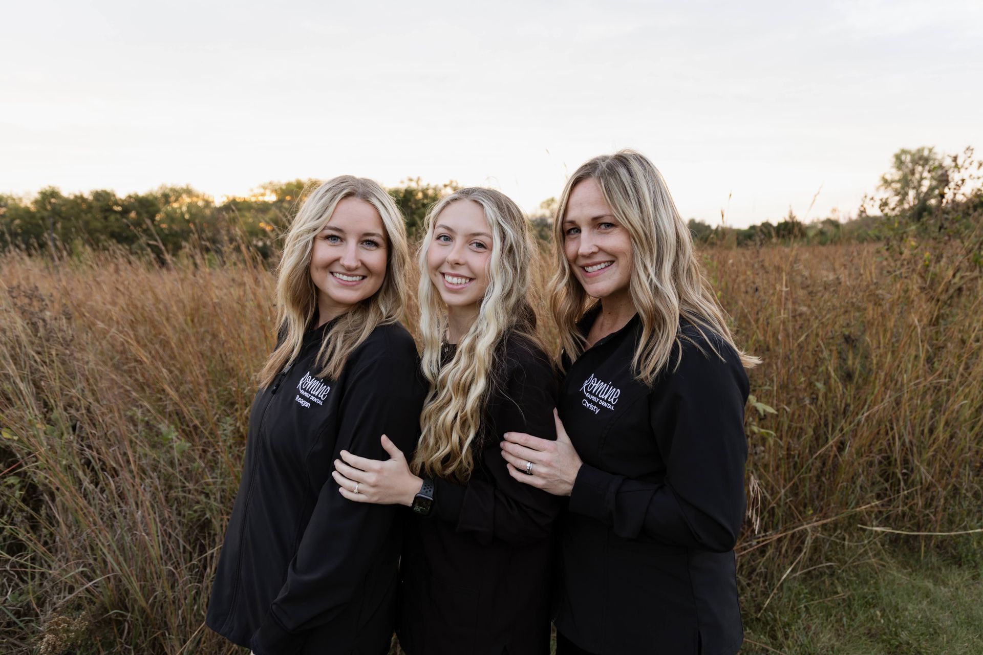 Three women in black shirts smiling, posing outdoors in a field of tall grass.